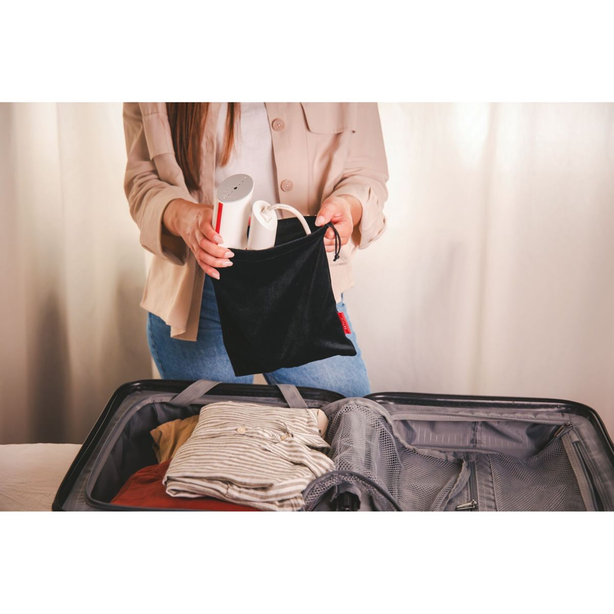 Eine Person packt einen Koffer. Sie hält eine schwarze Tasche mit weißen und roten Gegenständen über einem offenen Koffer mit Kleidung.