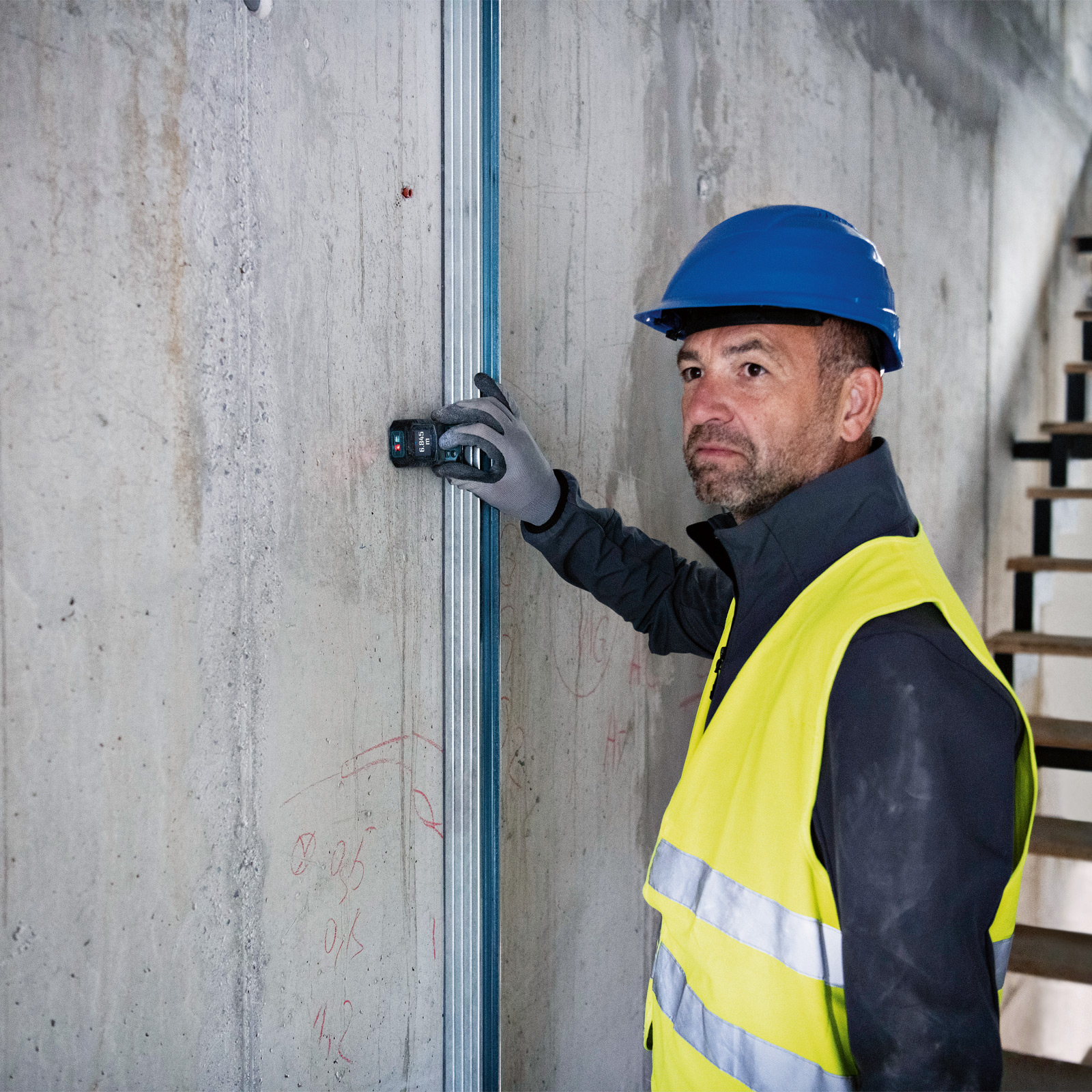 Ein Bauarbeiter mit blauem Helm und gelber Weste verwendet ein Messgerät an einer Betonmauer mit Metallstiften.