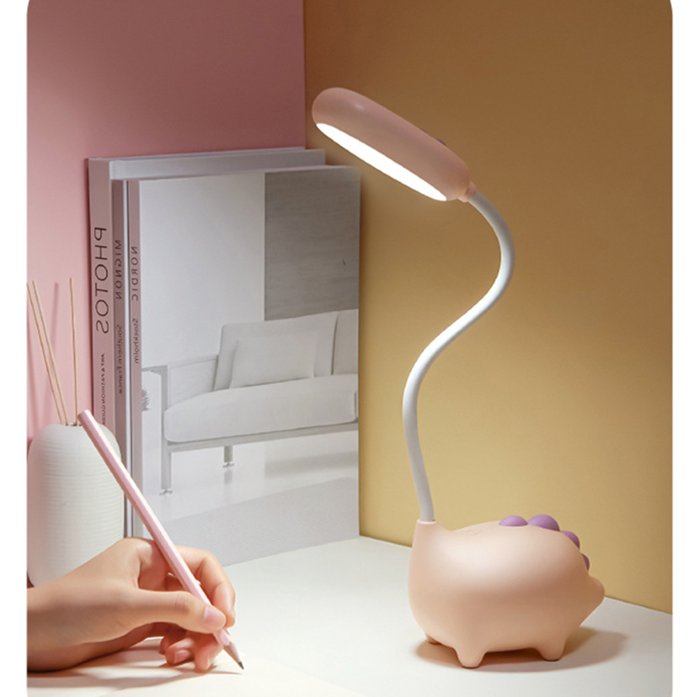Eine rosa Schreibtischlampe in Dinosaurierform. Eine Hand hält einen Bleistift und schreibt. Bücher und eine Vase mit Stöcken sind im Hintergrund.