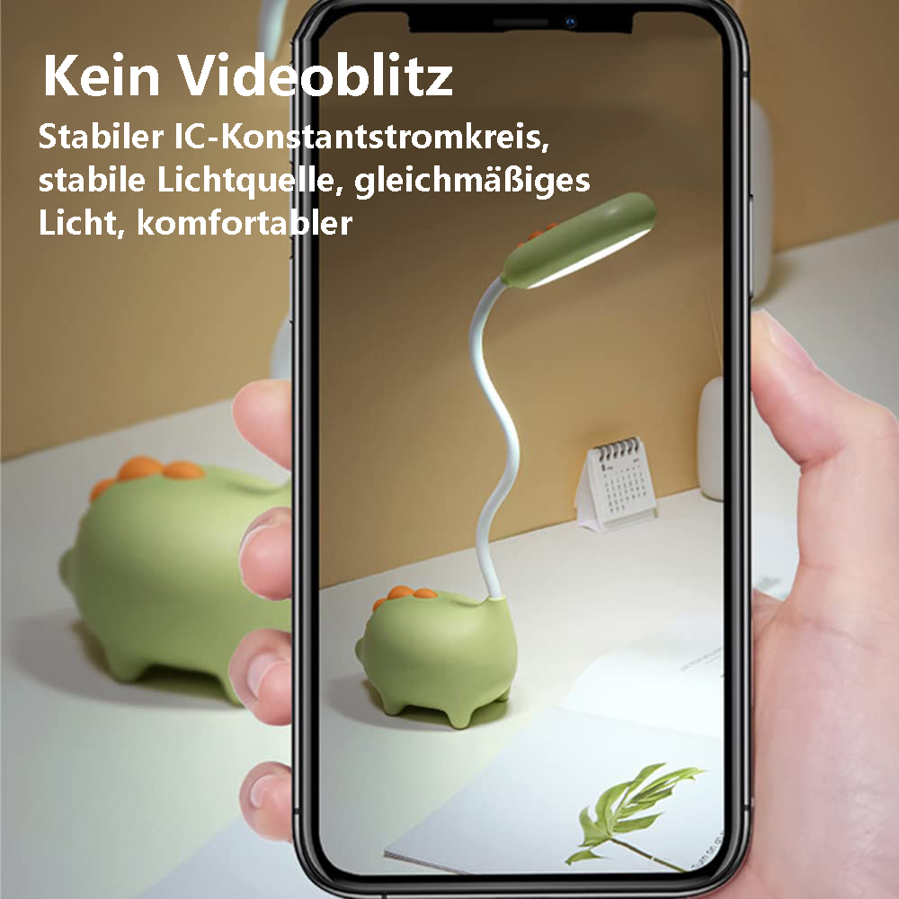 Ein Telefon zeigt eine grüne Dinosaurierlampe. Die Lampe steht auf einem Schreibtisch mit Kalender und Pflanze. Eine Hand hält das Telefon.