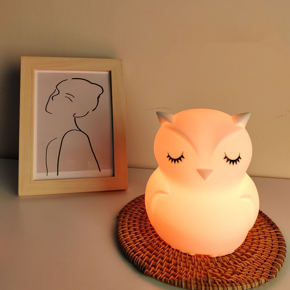 Eine orangefarbene, leuchtende Eulenlampe auf einer gewebten Matte. Links ein gerahmter Entwurf einer Frau.