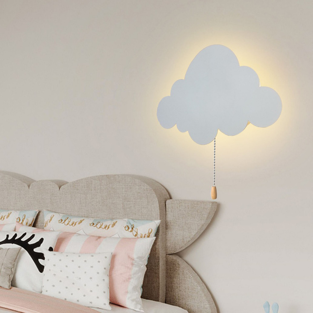 Eine wolkenförmige Wandlampe über einem Bett mit Kissen. Die Lampe hat eine Kette mit einem Holzgriff. Der Raum ist hell.
