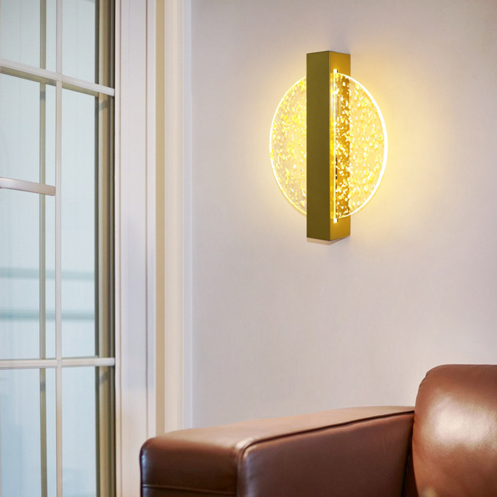 Eine Wandlampe mit goldenem Rahmen und kreisförmigem, beleuchtetem Glasdesign. Ein Fenster und ein Ledersofa sind ebenfalls sichtbar.