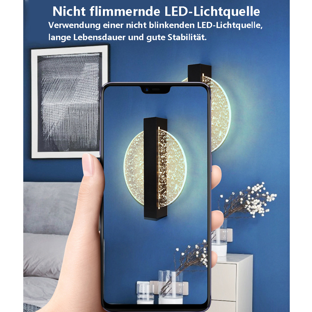 Eine Hand hält ein Telefon, das eine Wandlampe mit rundem Licht zeigt. Ein gerahmtes Bild und ein Nachttisch mit Deko befinden sich im Hintergrund.