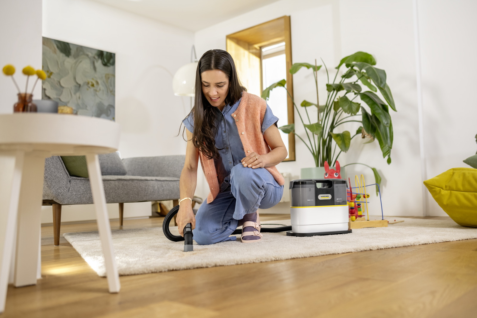 A woman vacuums a rug. She kneels, wearing a blue jumpsuit and orange vest. A Kärcher cleaner is on the rug. Plants and furniture are visible.