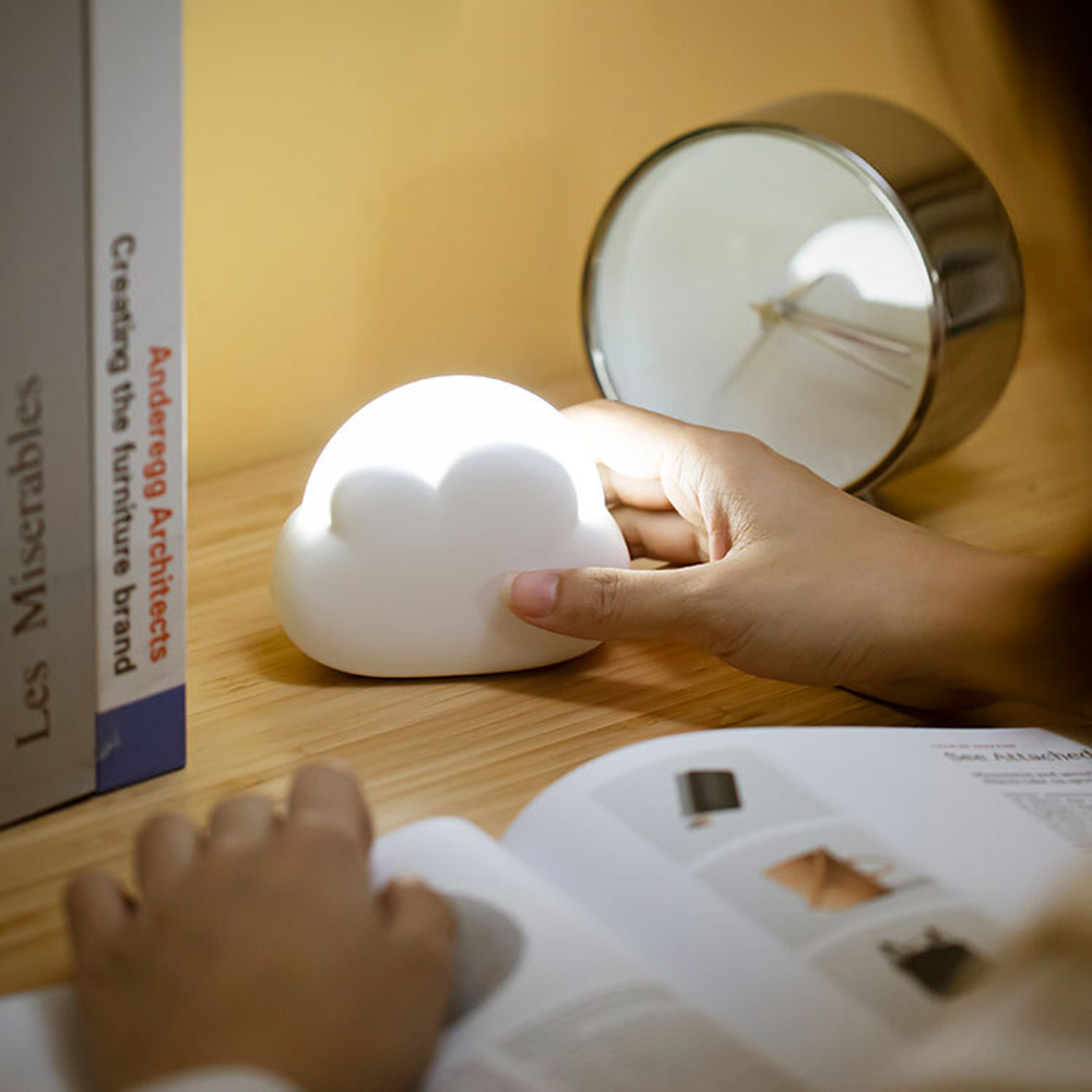 Eine Hand berührt eine wolkenförmige Lampe auf einem Holztisch. Ein offenes Buch, eine Uhr und Bücher sind ebenfalls vorhanden. Die Lampe leuchtet.