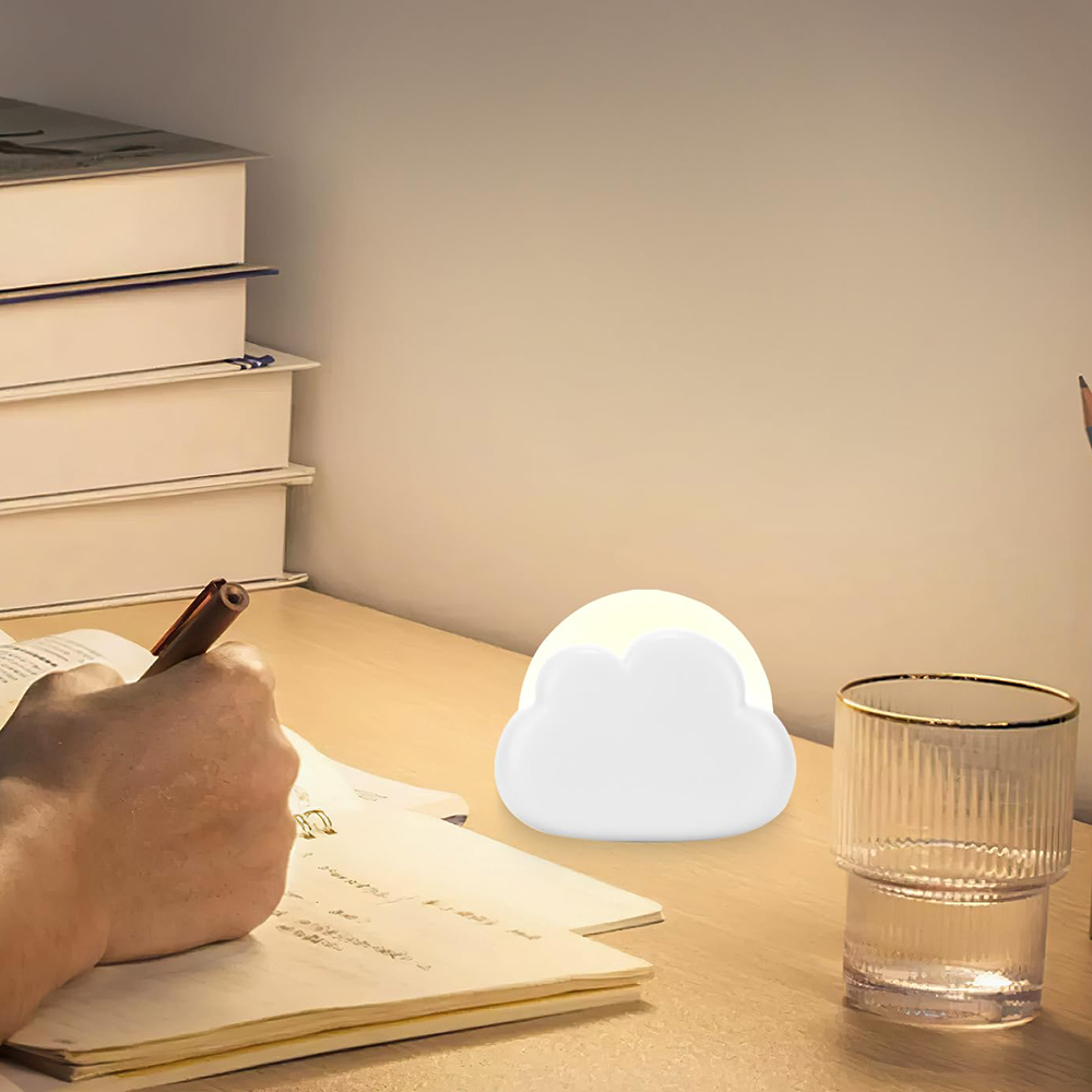 Eine Person schreibt mit einem Stift in ein Notizbuch. Eine wolkenförmige Lampe und ein Glas stehen auf dem Tisch.