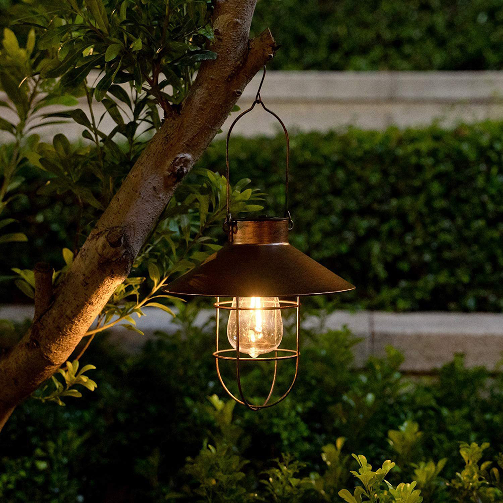 Eine Vintage-Außenlampe hängt an einem Ast. Sie hat einen dunklen Schirm und einen Käfig um eine Glühbirne. Grünes Laub ist im Hintergrund.
