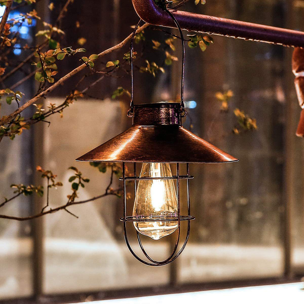 Eine kupferfarbene Lampe im Vintage-Stil hängt an einem Ast. Sie hat einen Käfig um eine leuchtende Glühbirne. Blätter und ein verschwommener Hintergrund sind ebenfalls vorhanden.