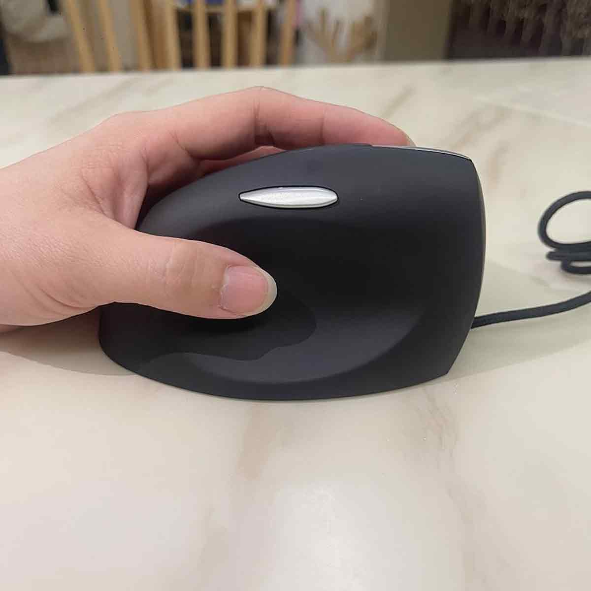 Eine Hand hält eine schwarze ergonomische Maus. Die Maus hat eine weiße Taste. Sie liegt auf einem hellen Tisch.