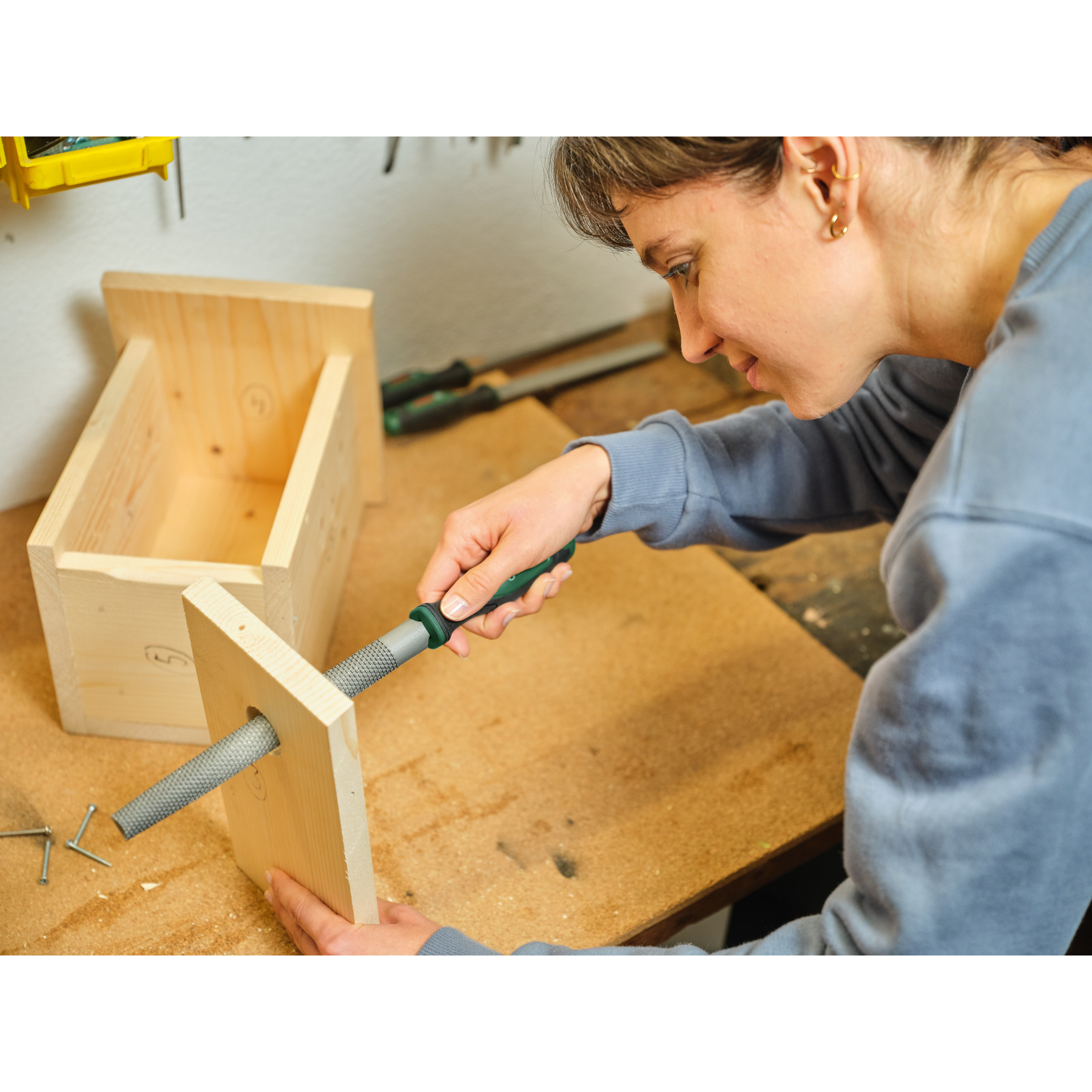 Eine Frau verwendet eine Raspel, um ein Holzbrett zu glätten. Ein Holzkasten befindet sich im Hintergrund.