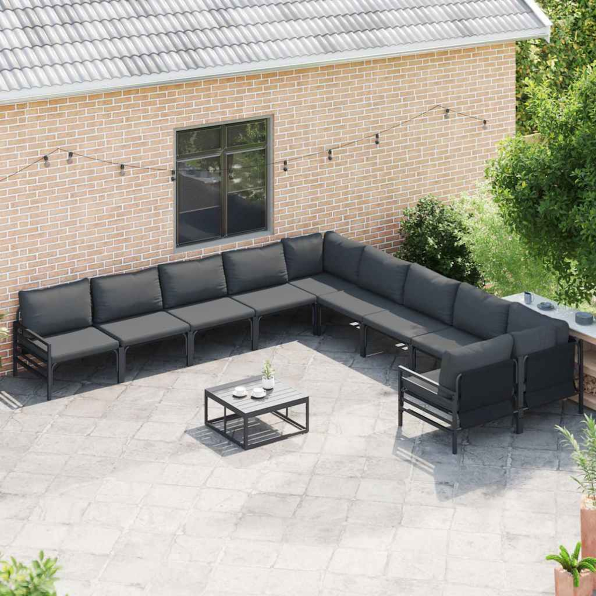 Patio esterno con divano grigio, tavolino con tazze, muro di mattoni, finestra e piante verdi.