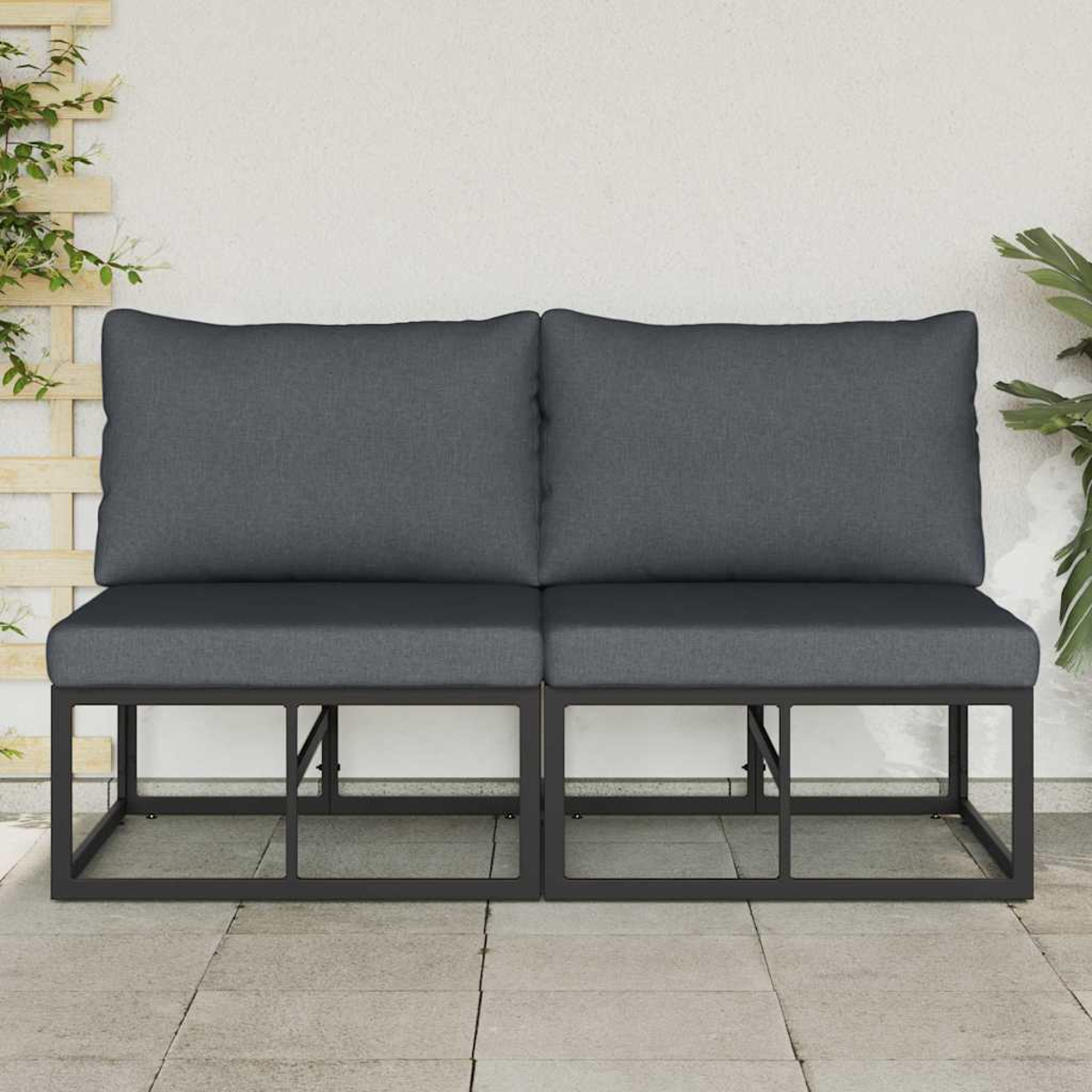 Dos asientos exteriores con cojines grises. Marcos de metal negro. Fondo de pared blanca. Plantas a la derecha e izquierda. Suelo de baldosas de piedra.