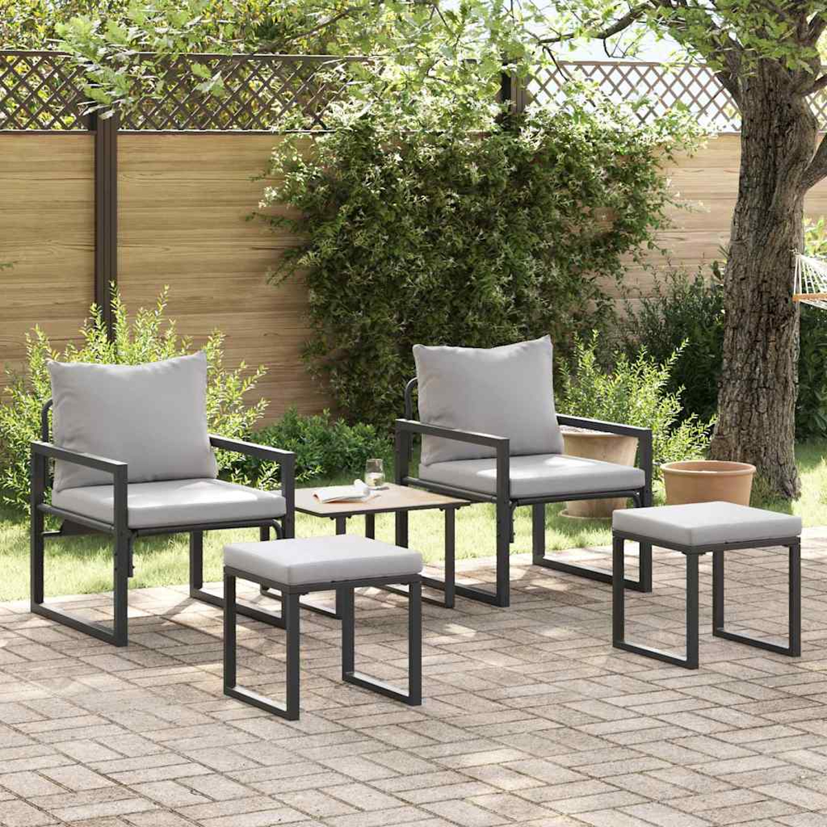 Szene im Freien mit zwei Stühlen, zwei Hockern und einem kleinen Tisch. Graue Kissen auf schwarzen Metallrahmen. Ziegelterrasse mit grünem Hintergrund.