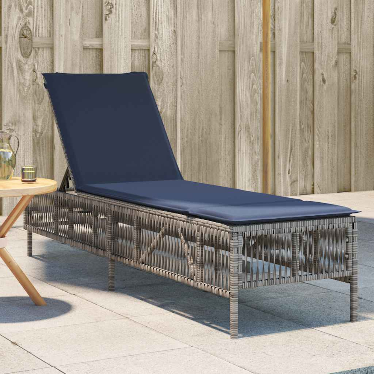 Eine Korbliege mit blauem Kissen steht auf einer Terrasse. Ein kleiner Tisch mit einem Glas und ein Holzzaun sind im Hintergrund.