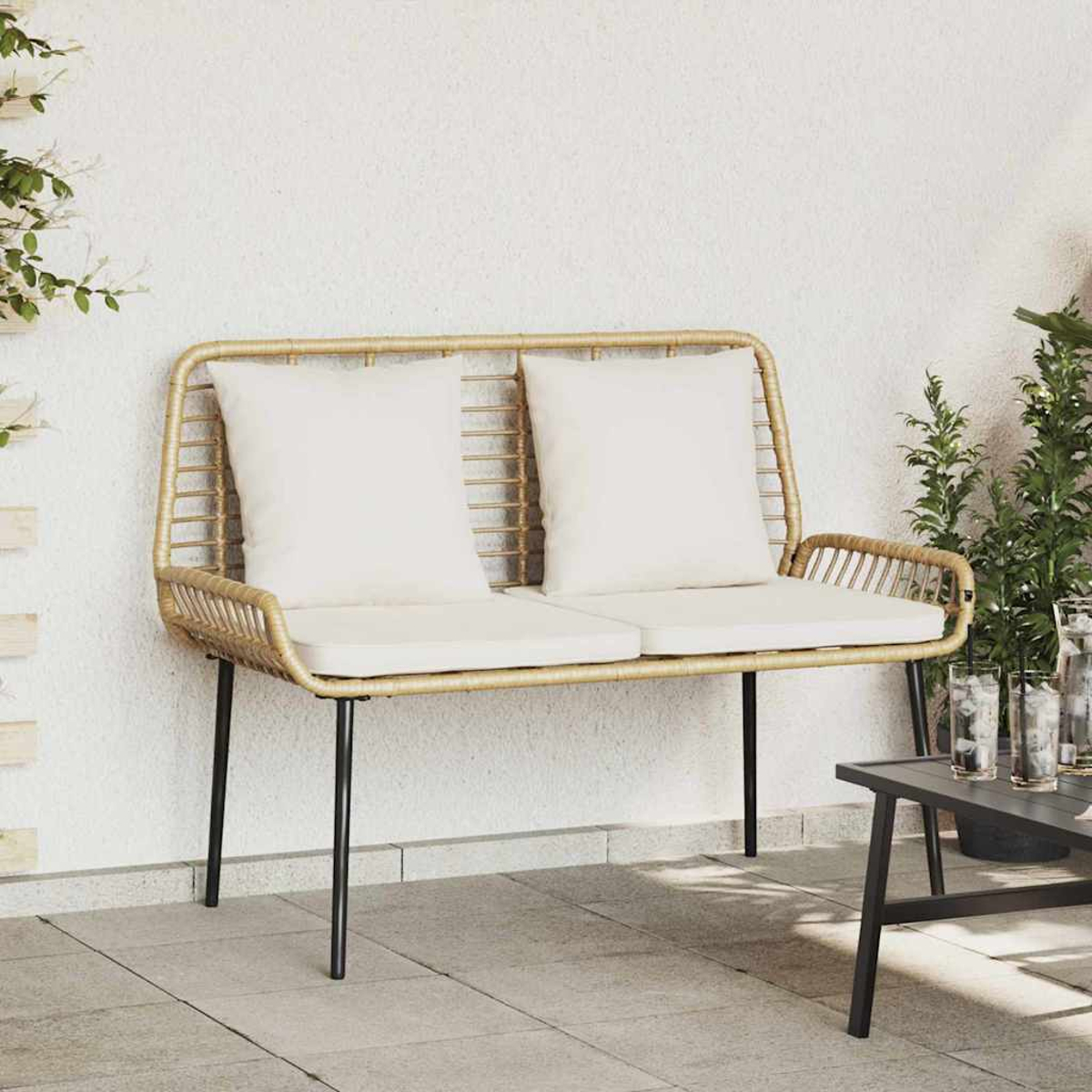 Eine Korbsitzbank mit weißen Kissen steht auf einer Terrasse. Grüne Pflanzen und ein Tisch mit Gläsern sind im Hintergrund.