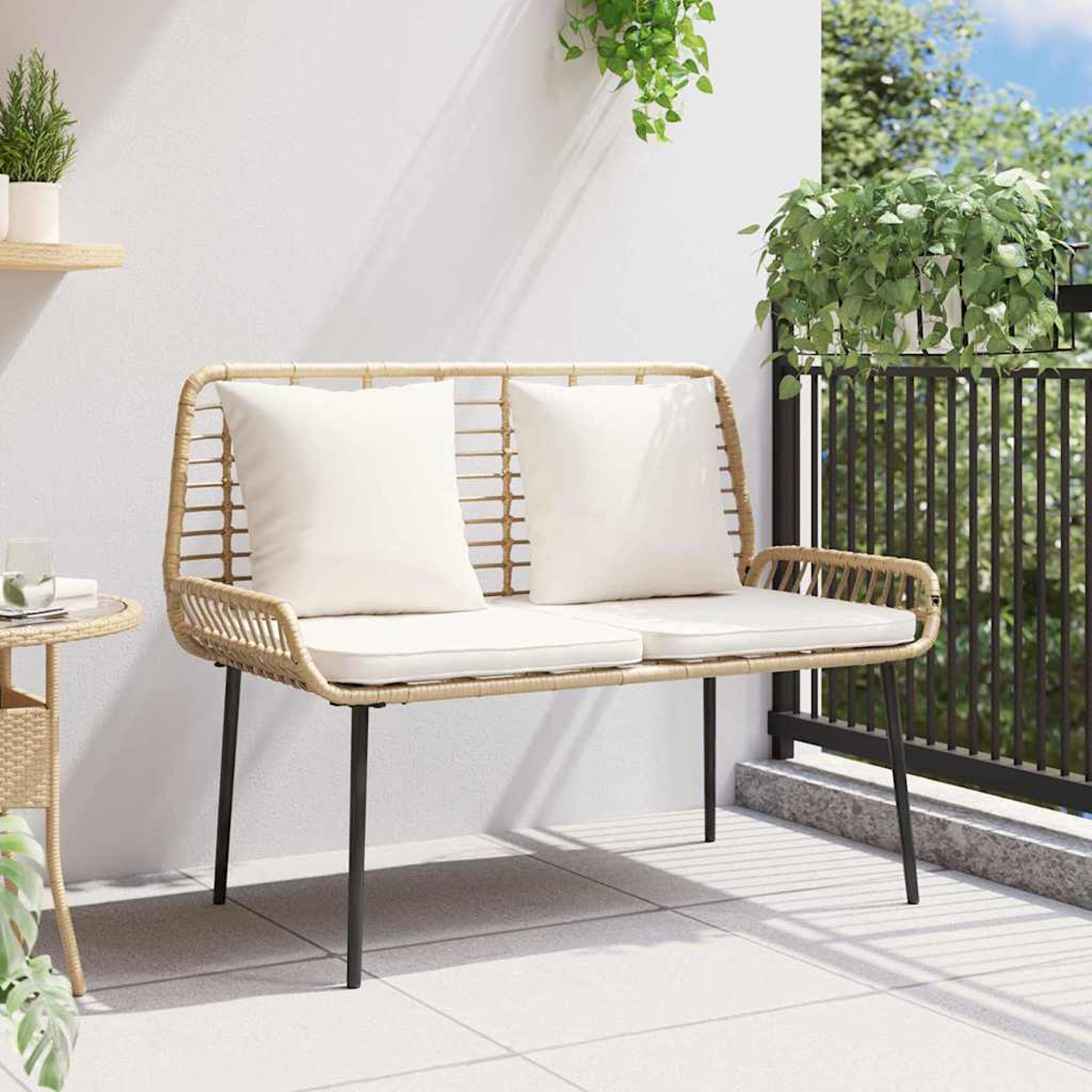 Eine Korbsitzbank mit weißen Kissen steht auf einem Balkon. Pflanzen hängen im Hintergrund. Links ist ein kleiner Tisch.