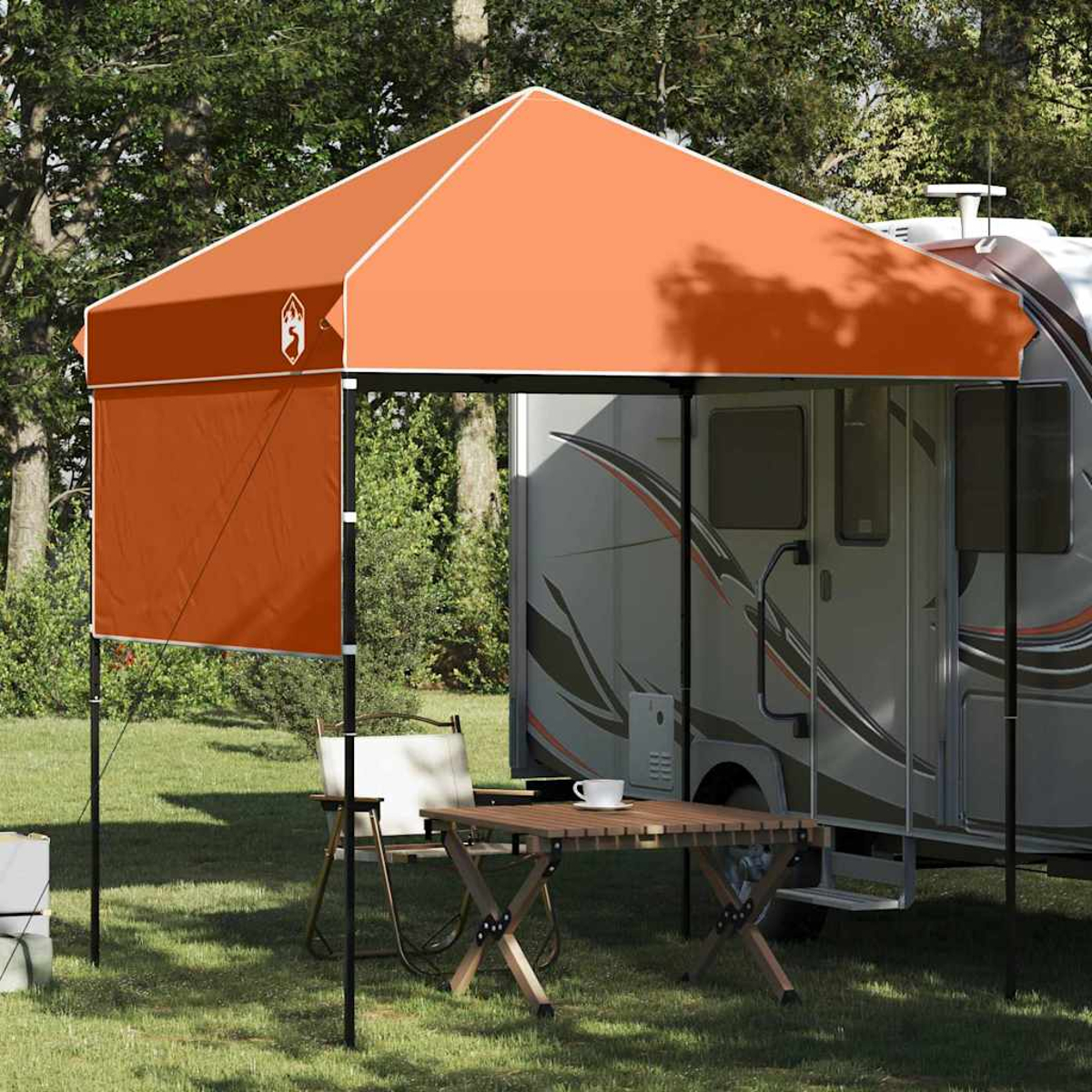 Una tenda arancione con bordo bianco è accanto a un camper. Un tavolo e una sedia sono disposti davanti. Alberi visibili sullo sfondo.
