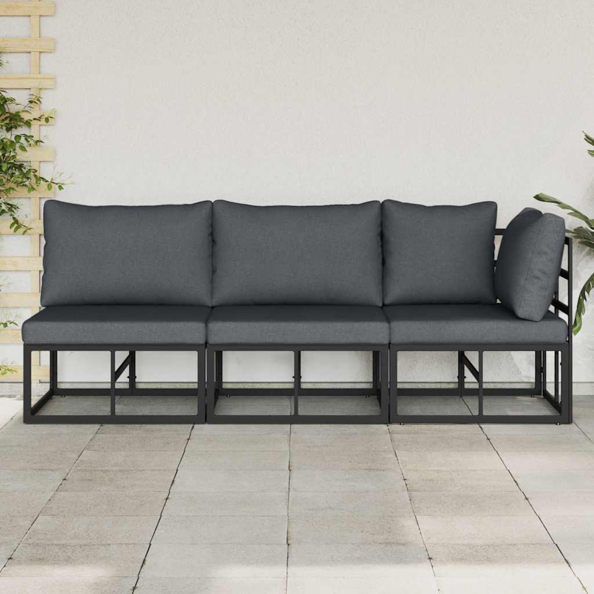 Un divano da esterno a tre pezzi con cuscini grigi e struttura in metallo nero. È posizionato su un patio piastrellato, con un muro bianco sullo sfondo.
