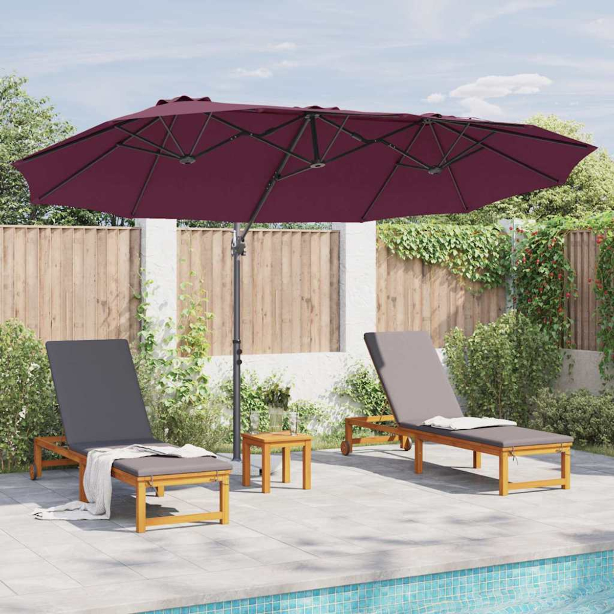 Due sedie a sdraio sotto un grande ombrello bordeaux. Un tavolino tra di loro. Fogliame verde sullo sfondo, piscina in primo piano.