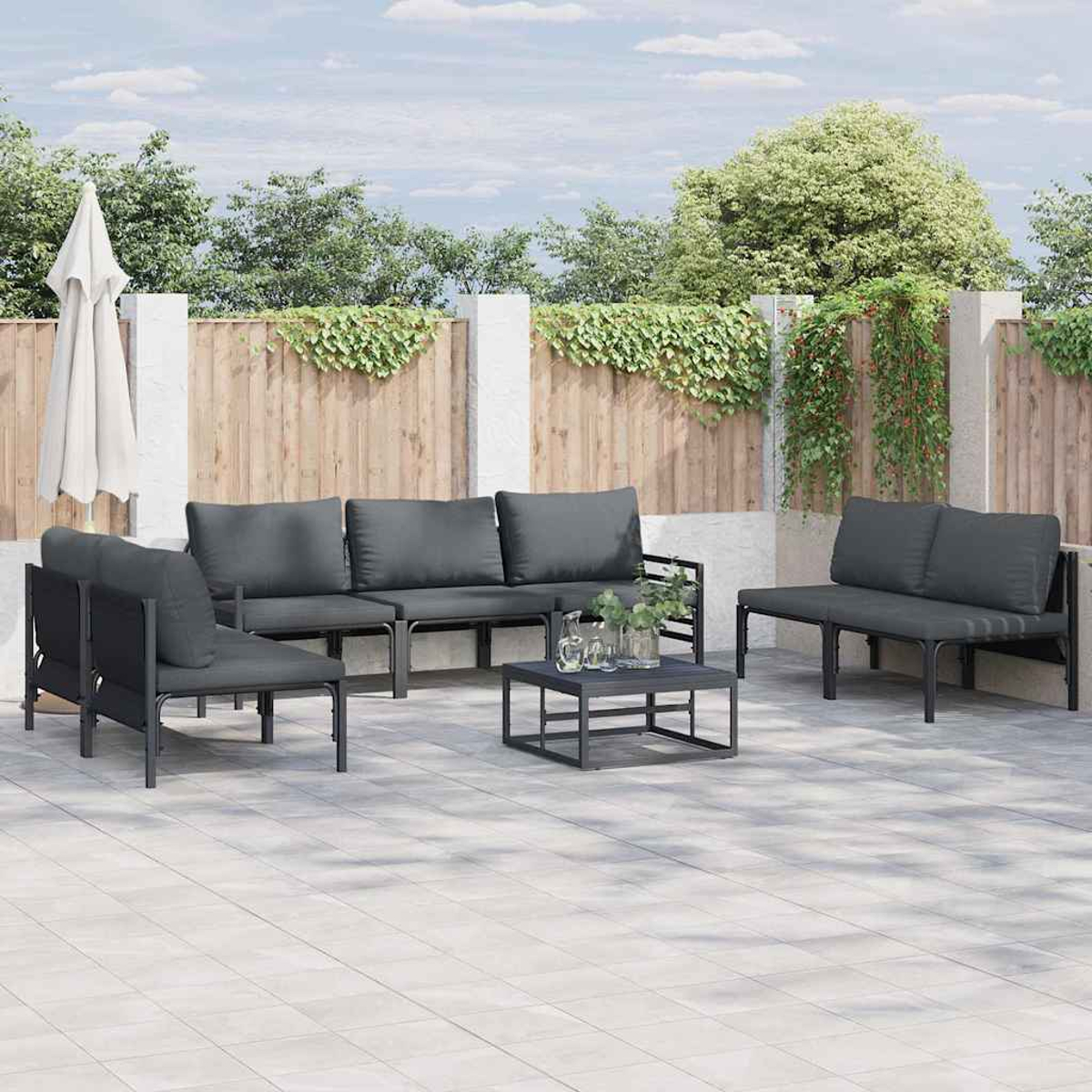 Juego de patio exterior con cojines grises. Incluye sofá, sillas y mesa de centro. Fondo: valla de madera y vegetación.