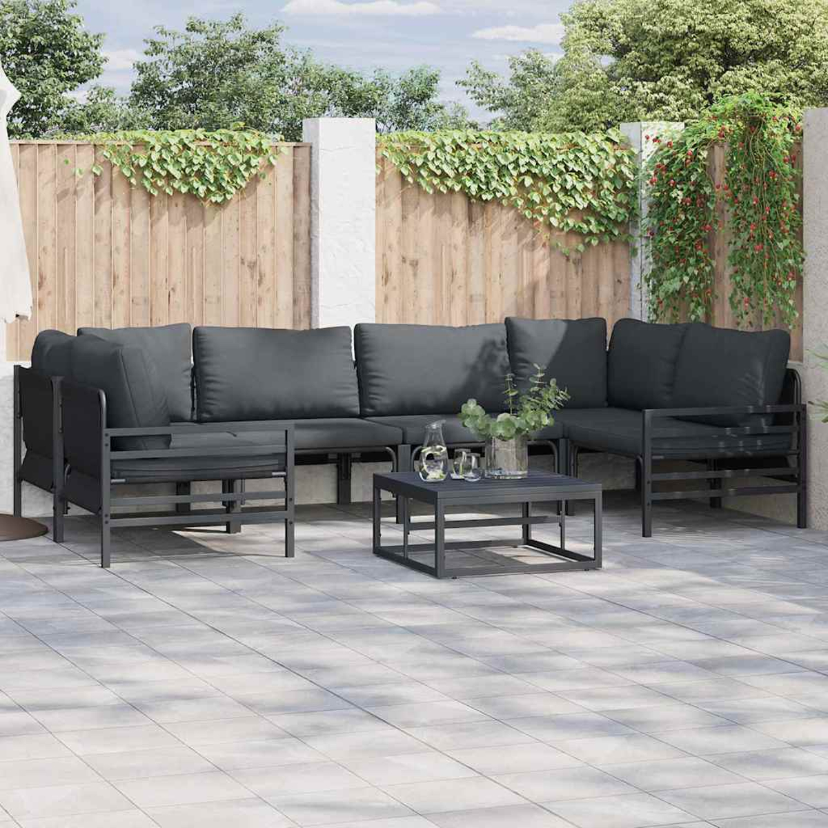 Garten-Sitzgruppe. Dunkelgraues Ecksofa, passender Couchtisch und Kissen. Hintergrund Holzzaun mit Grün.
