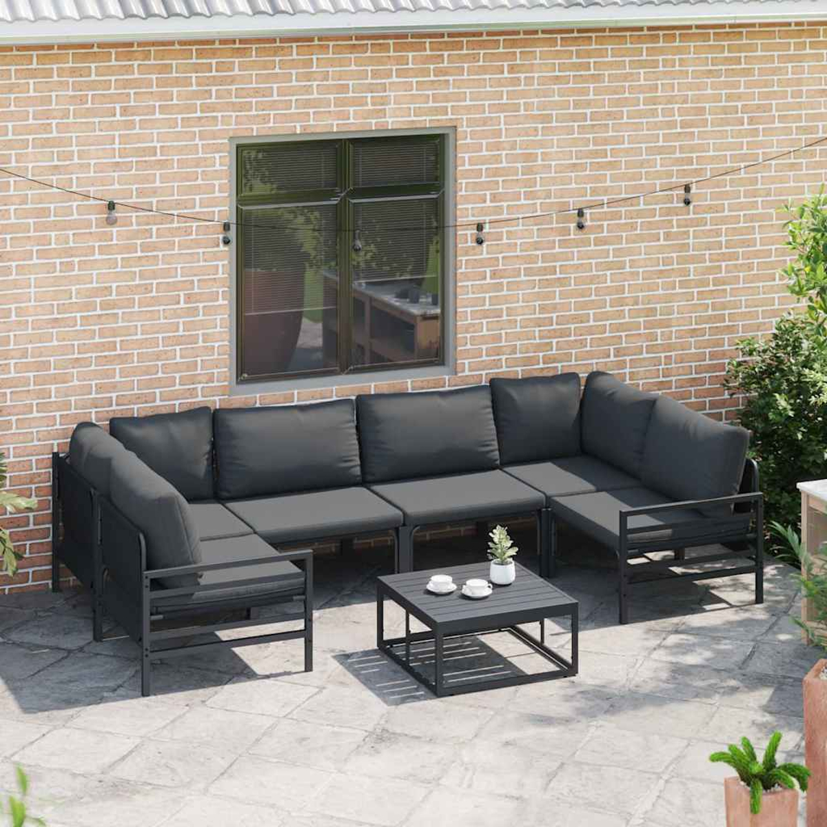 Gartencouch mit grauen Kissen und einem passenden Couchtisch auf einer Terrasse. Ziegelmauer und Fenster im Hintergrund.