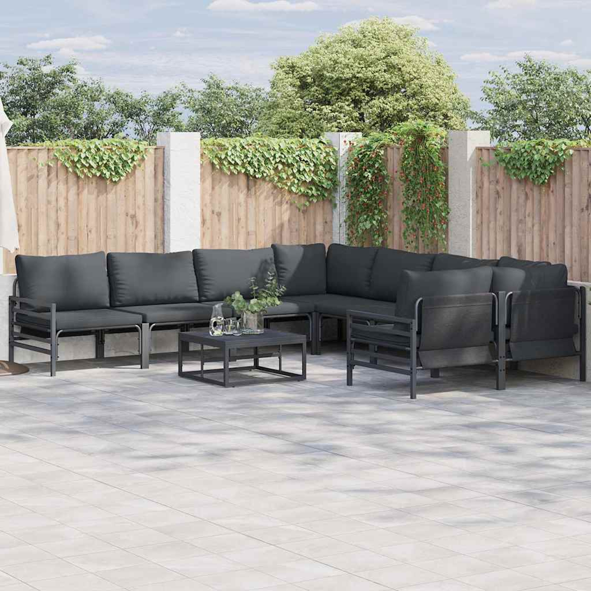 Set da patio all'aperto con cuscini grigio scuro. Un tavolino è di fronte. Recinzione in legno sullo sfondo, con vegetazione e alberi.