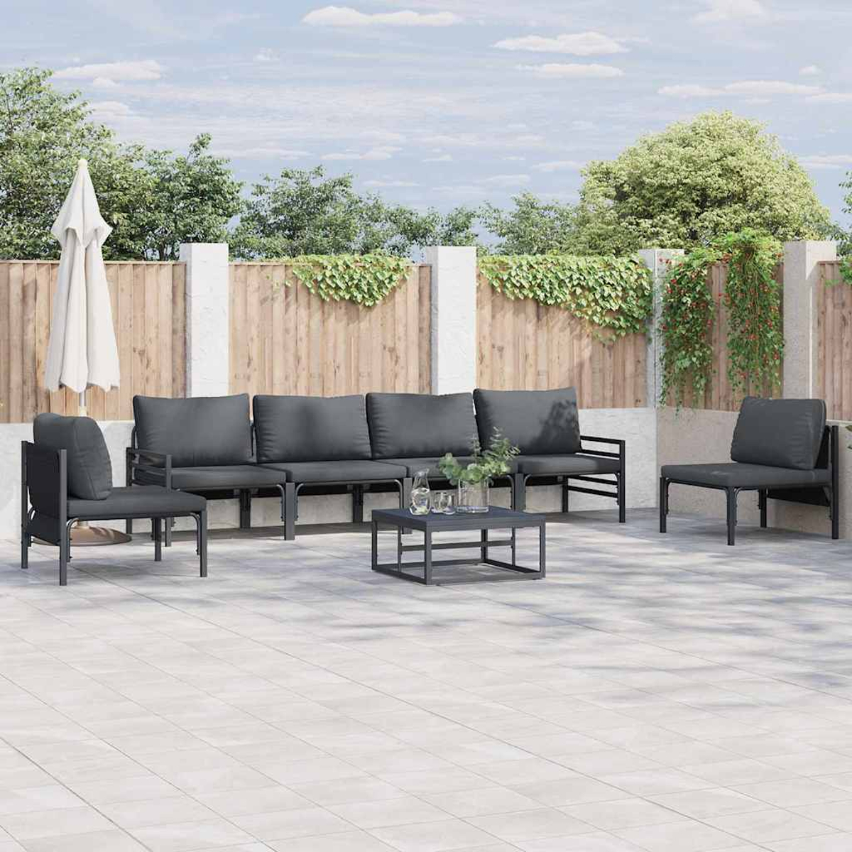 Set da patio esterno: divani, sedie e tavolo grigi su un patio in pietra. Ombrellone bianco e recinzione in legno con verde sullo sfondo.