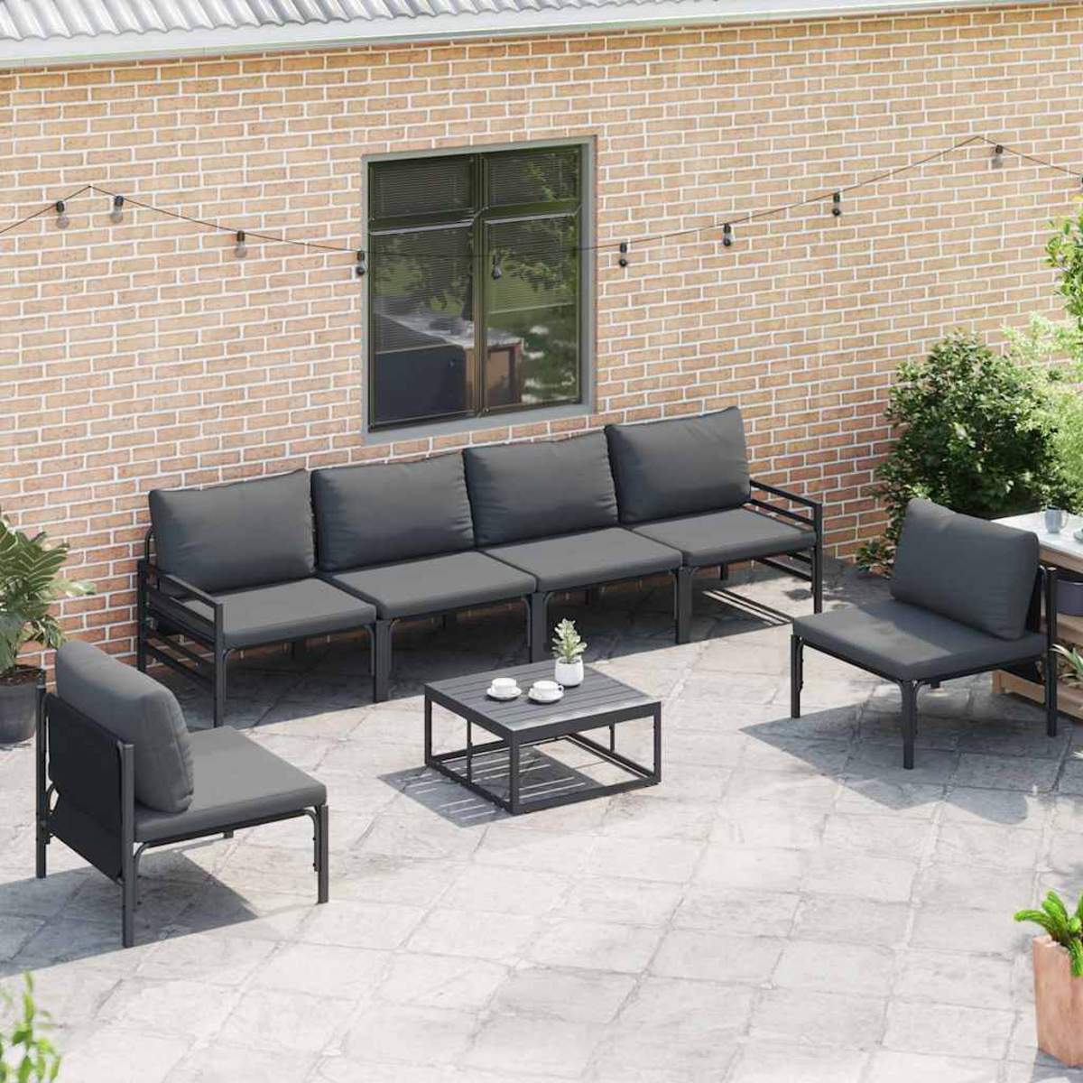 Set di mobili da patio con cuscini grigi. Include un divano, due sedie e un tavolino. Sfondo muro di mattoni.