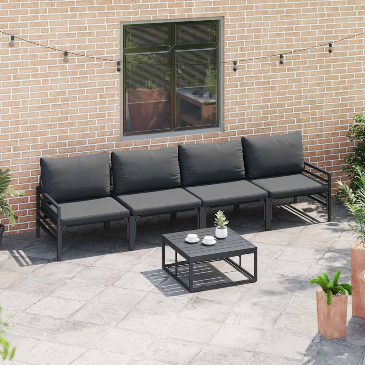 Outdoor-Terrasseneinrichtung. Graues Viersitzer-Sofa, kleiner Tisch mit Tassen, Backsteinwand im Hintergrund, Fenster und Topfpflanzen.