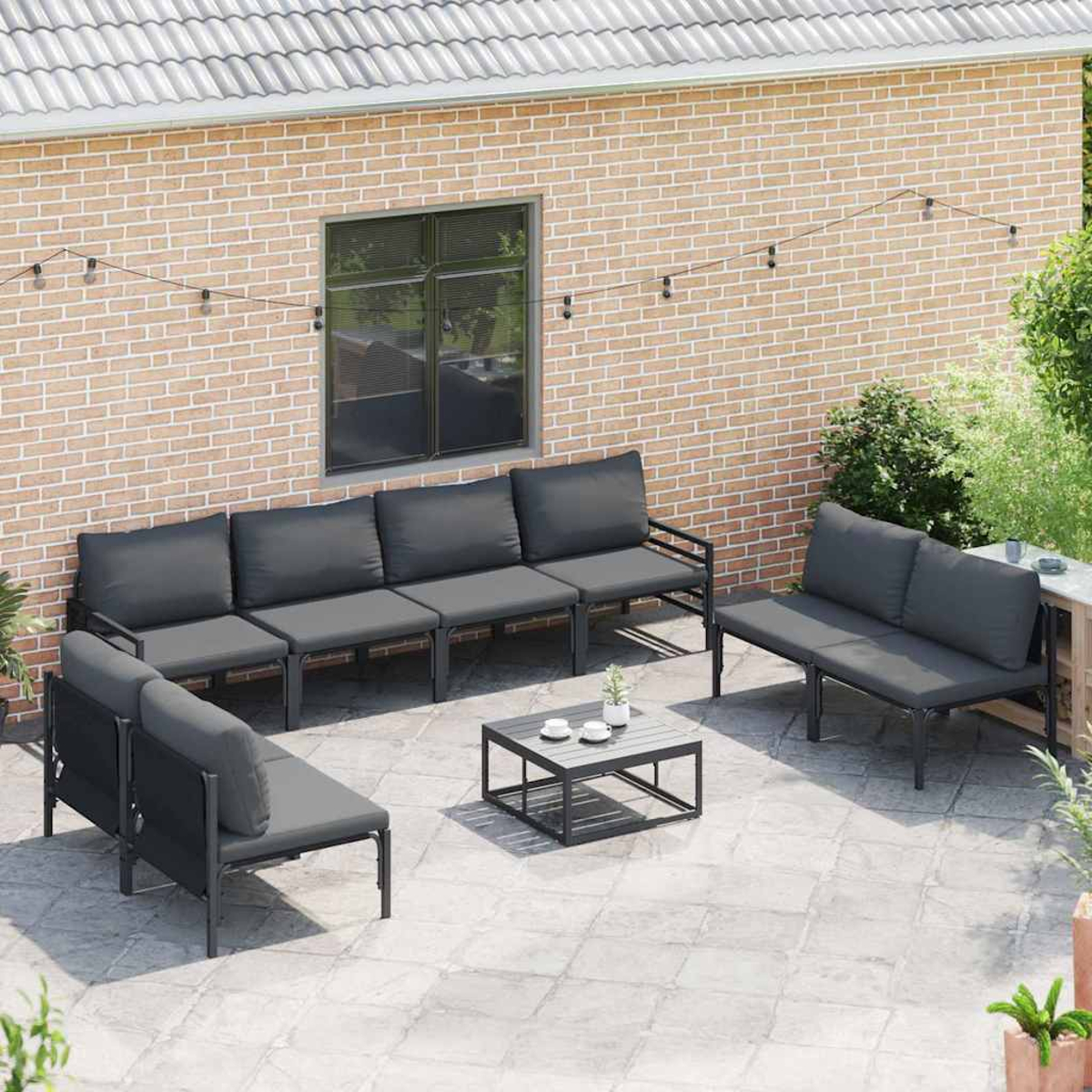 Terrassenset mit grauen Kissen. Beinhaltet ein Sofa, eine Doppelsitzbank und einen Couchtisch. Der Hintergrund ist eine Backsteinmauer und Grün.