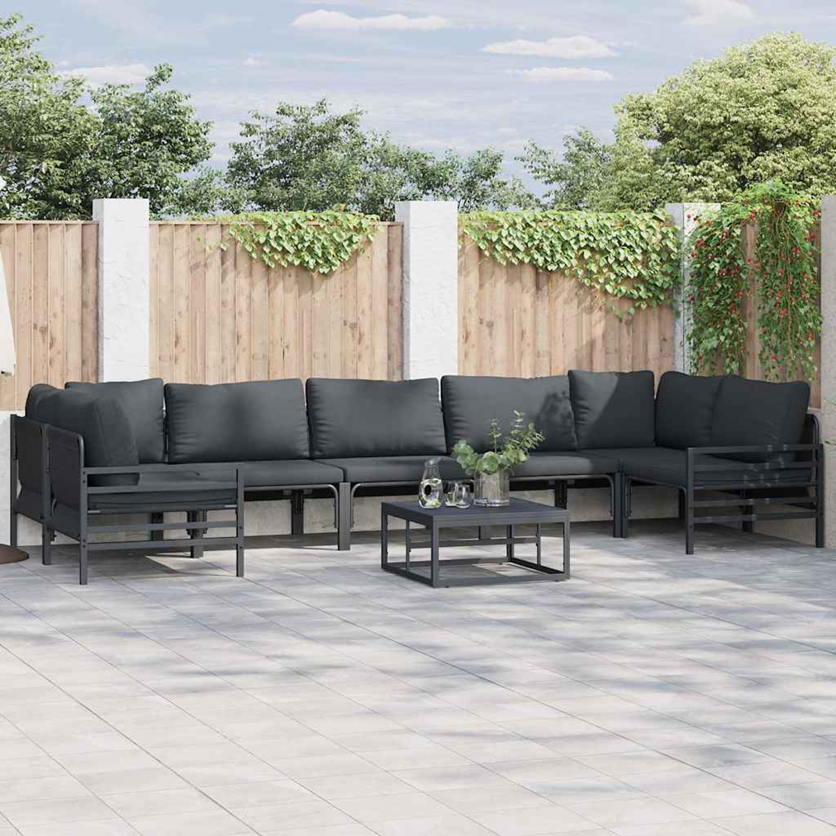 Szene im Freien mit dunkelgrauem Ecksofa, kleinem Tisch und Holzzaun mit Grün. Sonniger Tag, Terrasse.