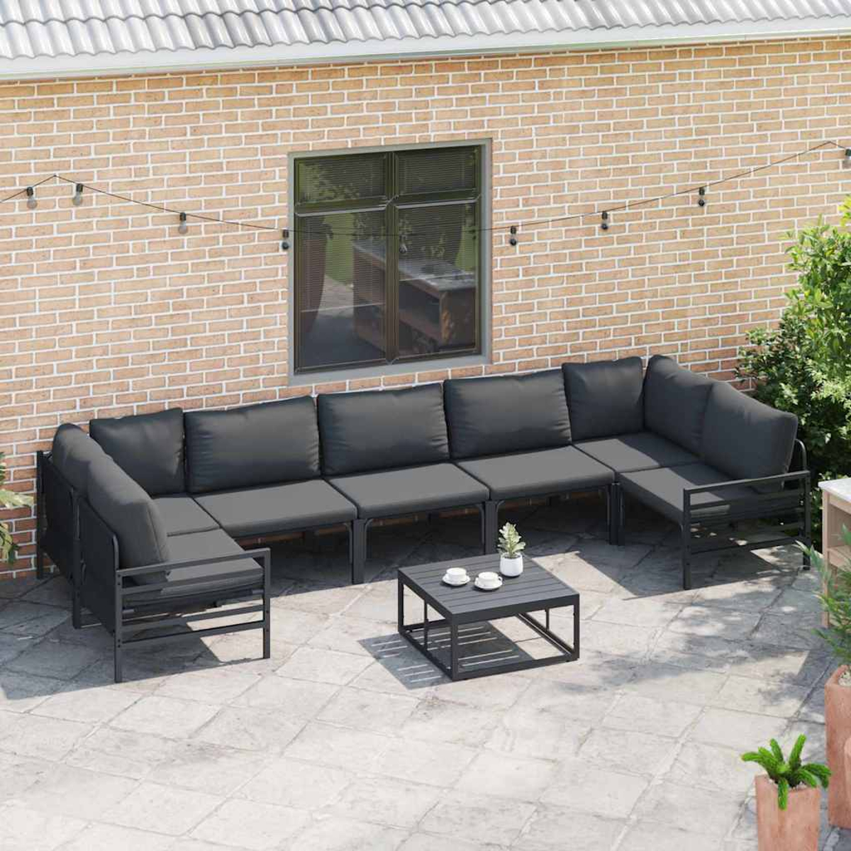 Eine Outdoor-Terrasseneinrichtung mit einem grauen Ecksofa und einem quadratischen Couchtisch. Eine Backsteinmauer und Lichterketten vervollständigen die Szene.