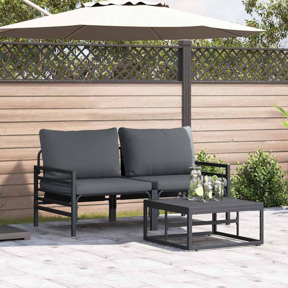 Set da patio all'aperto con cuscini grigi, struttura in metallo nero e tavolino. Un ombrello bianco e una recinzione in legno sullo sfondo.