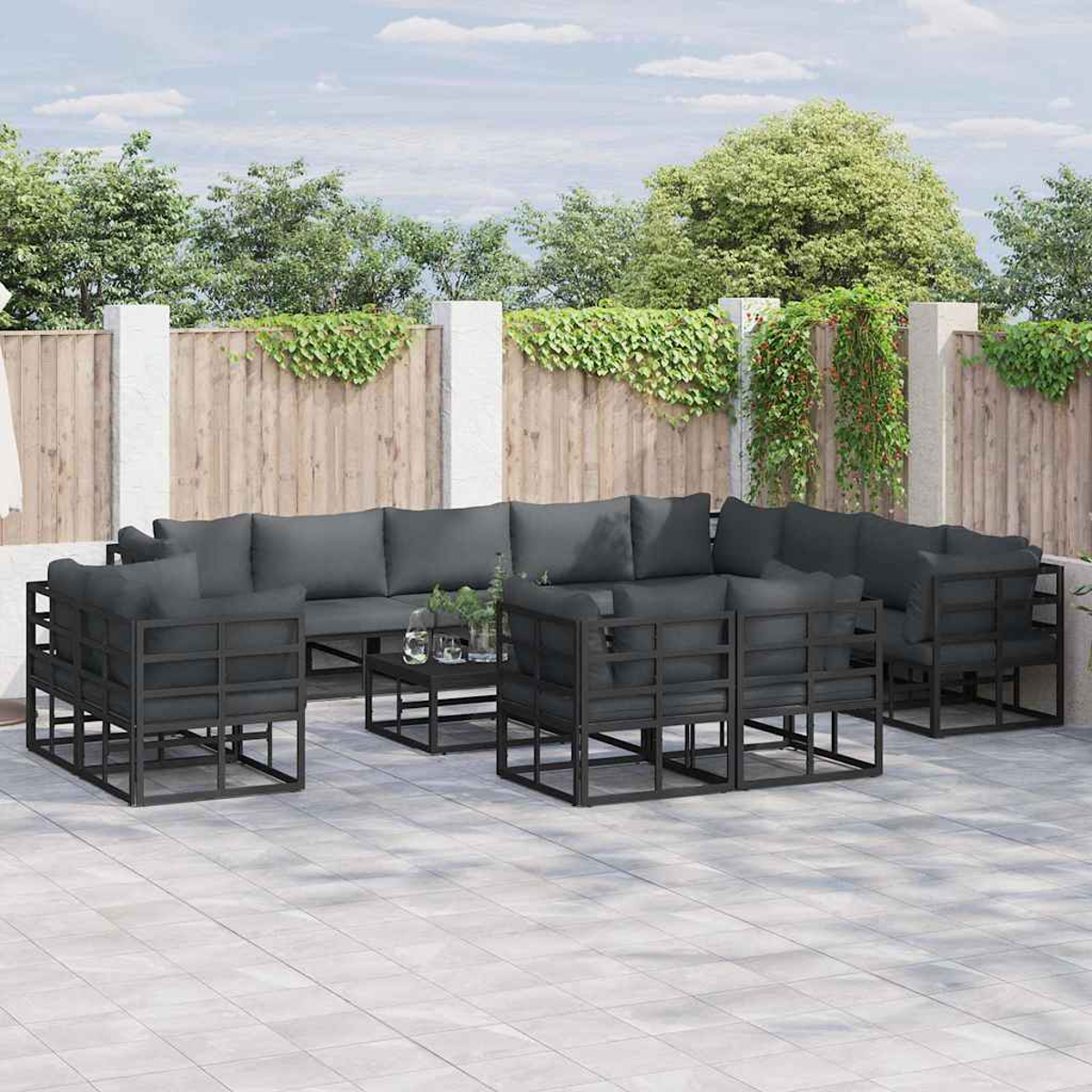 Set di mobili da patio con cuscini grigi. Include divani, poltrone e un tavolo. Sullo sfondo una recinzione in legno e vegetazione.
