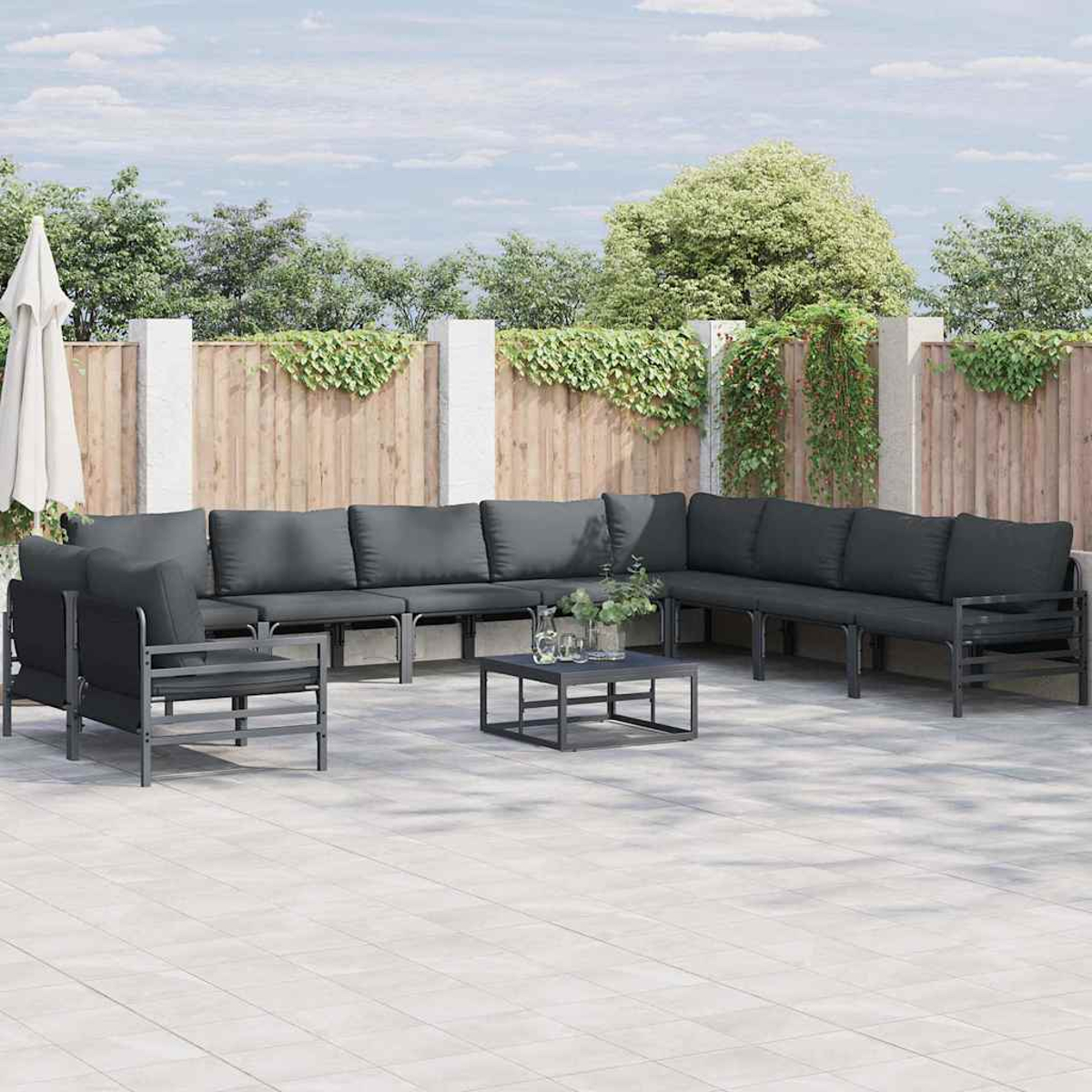 Outdoor-Sitzbereich mit großem dunkelgrauen Ecksofa, kleinem Tisch und Sonnenschirm vor Holzzaun.