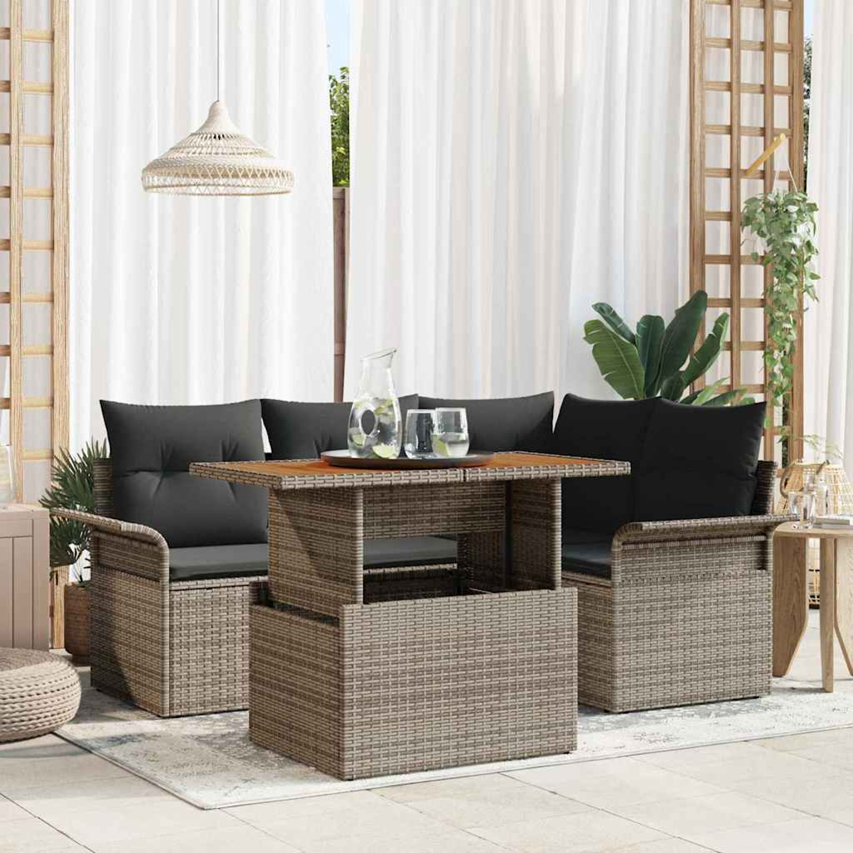 Garten-Set mit gewebten Möbeln, schwarzen Kissen und Holztisch. Weiße Vorhänge im Hintergrund. Pflanzen und Leuchte sind sichtbar.