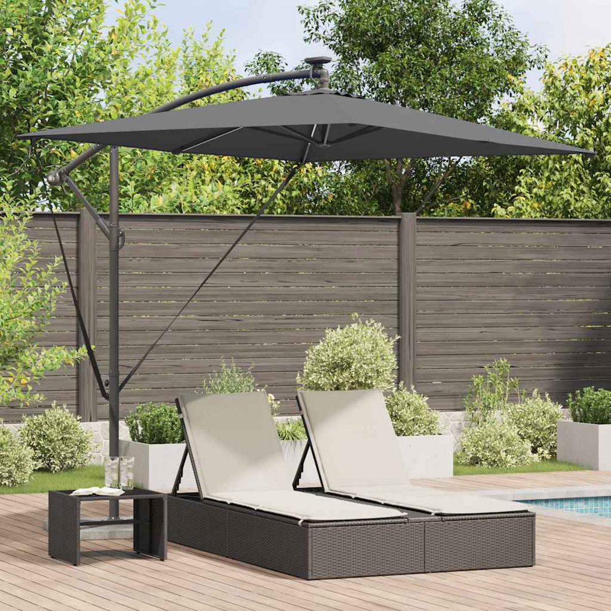 Scena all'aperto con due sedie a sdraio, un tavolino e un grande ombrello grigio. Un ponte di legno è di fronte a una piscina e a una recinzione.