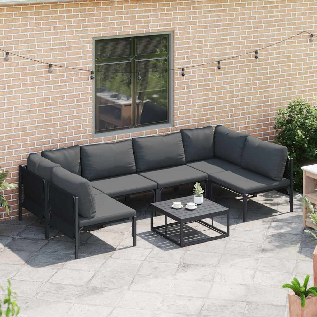 Divano componibile da esterno con cuscini grigi su un patio. Include un tavolo con tazze da caffè e un muro di mattoni sullo sfondo.
