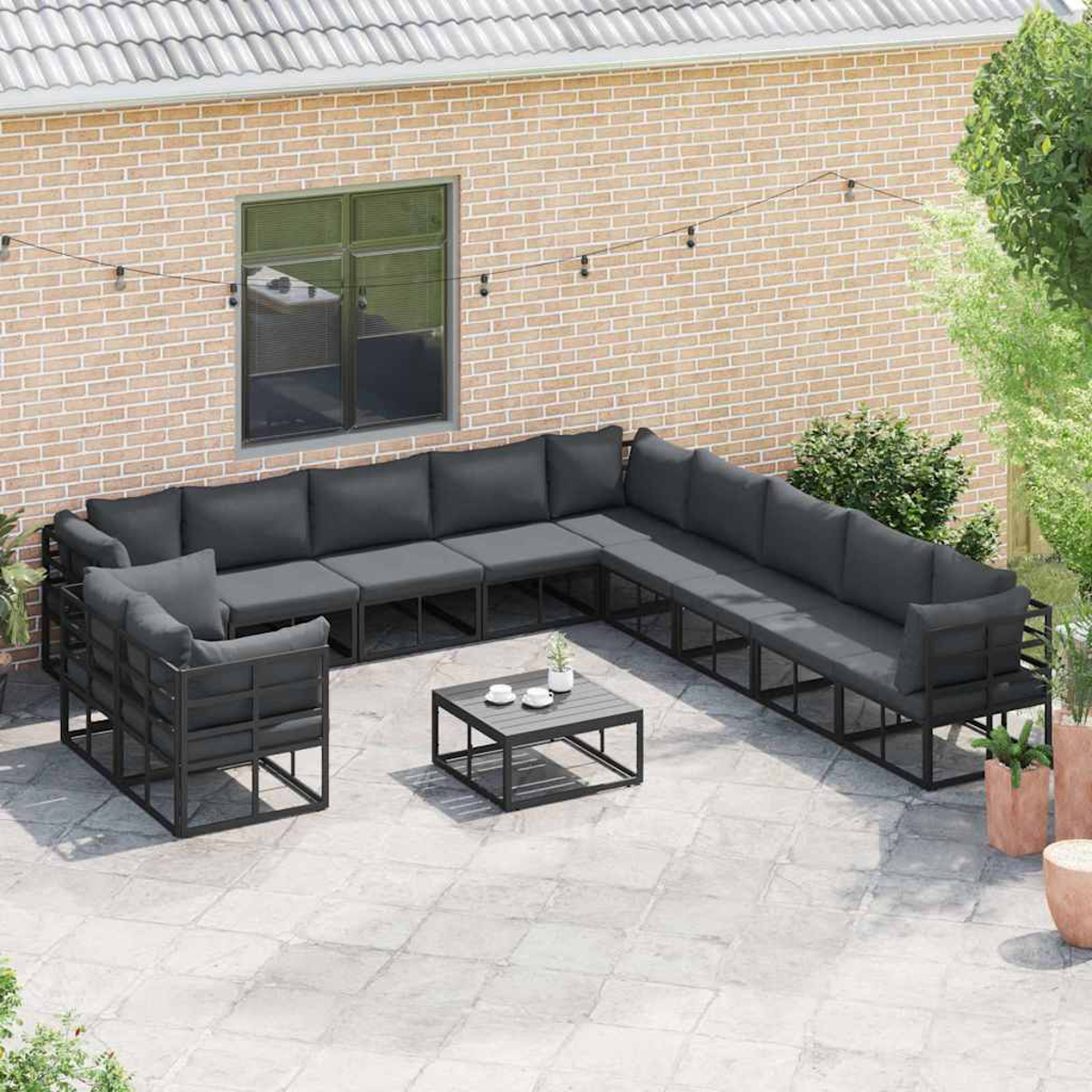 Terrassen-Set mit grauen Kissen, schwarzem Rahmen und kleinem Tisch. Backsteinmauer und Grün in der Hintergrund. Lichterketten darüber.