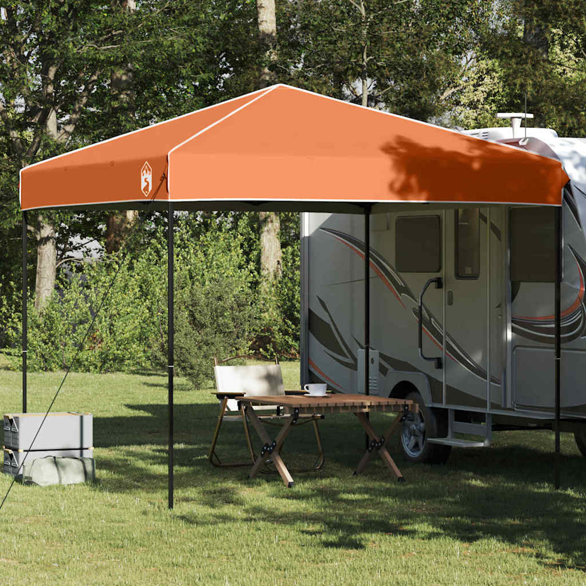 Ein oranger Baldachin beschattet einen Campingbereich mit Tisch, Stühlen und einem Wohnwagen. Bäume sind im Hintergrund.