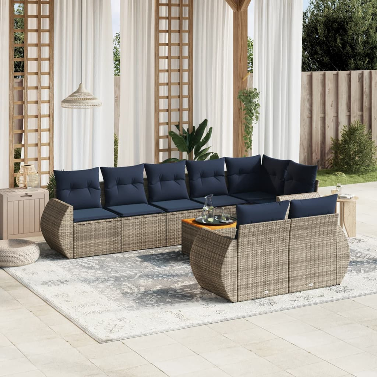 Outdoor-Sitzgruppe mit blauen Kissen auf einem gemusterten Teppich, unter einer Pergola und transparenten Vorhängen. Ein Tisch mit Getränken steht im Zentrum.