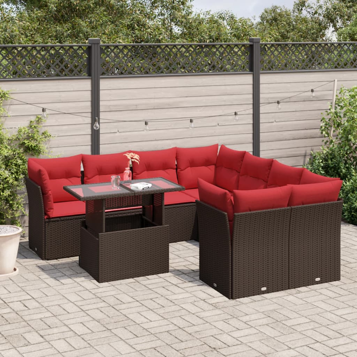 Terrassenmöbel-Set mit roten Kissen und dunkelbraunem Korbgeflecht. Enthält einen Tisch und einen Zaun im Hintergrund. Gepflasterter Boden.