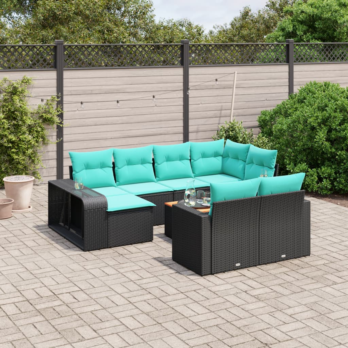 Ein schwarzes Outdoor-Sofa aus Korbgeflecht mit türkisfarbenen Kissen steht auf einer Terrasse. Ein Zaun und Grünzeug sind im Hintergrund.
