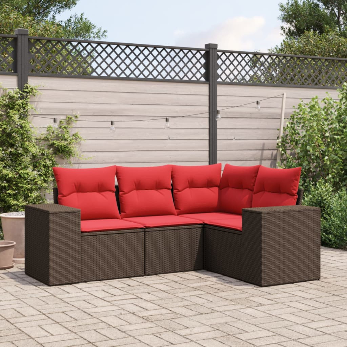 Ein Ecksofa im Freien mit roten Kissen. Es ist aus braunem Korbgeflecht und steht auf einer Ziegelterrasse. Ein Zaun und Grünzeug sind im Hintergrund.