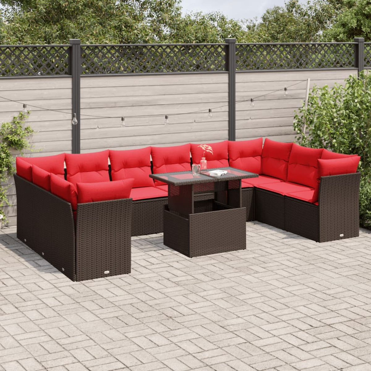 Seccional de mimbre para exteriores con cojines rojos y mesa a juego en un patio. Fondo con valla y vegetación.