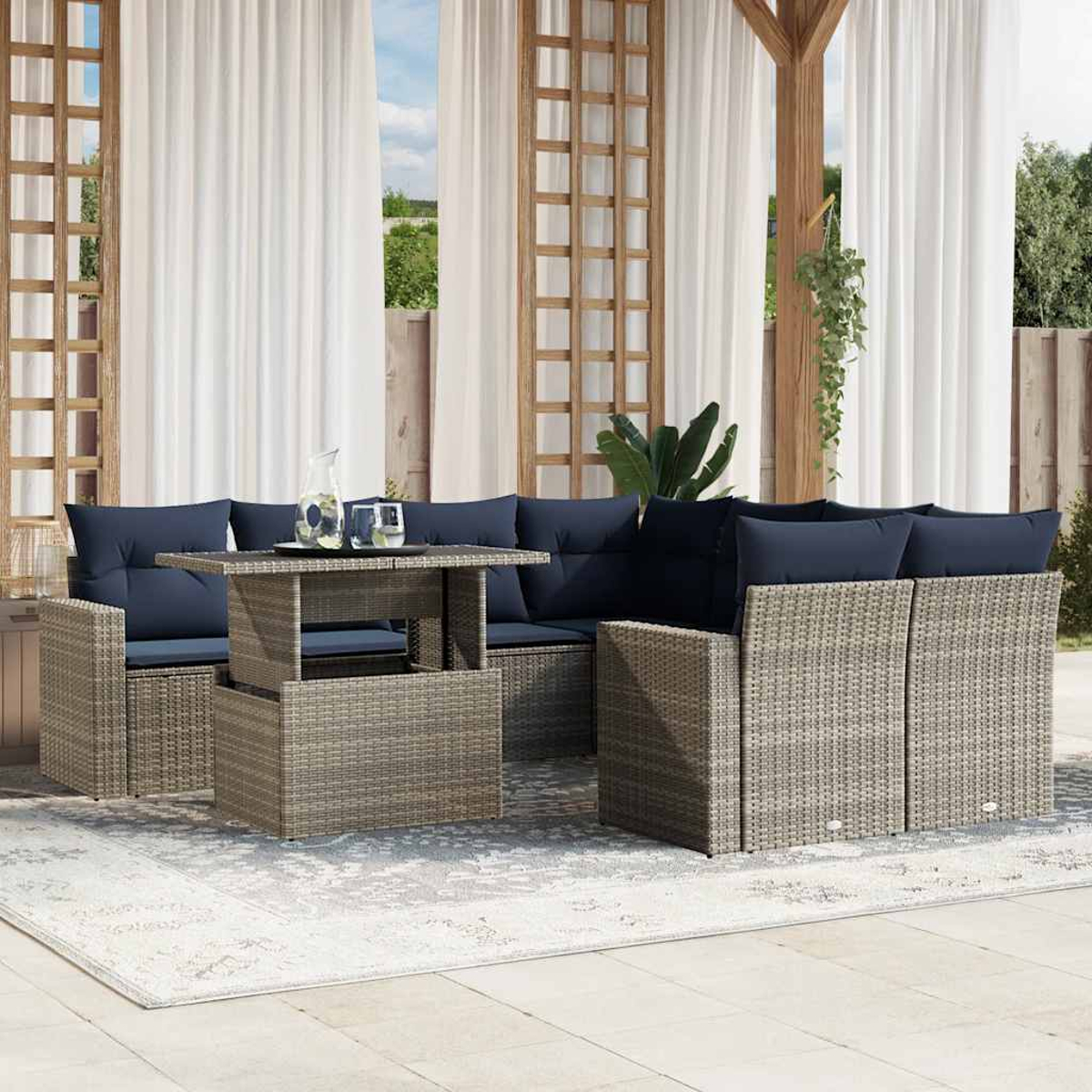 Terrassengarnitur mit grauen Korbmöbeln, marineblauen Kissen und einem Tisch mit Hubfunktion. Weiße Vorhänge und Grün im Hintergrund.