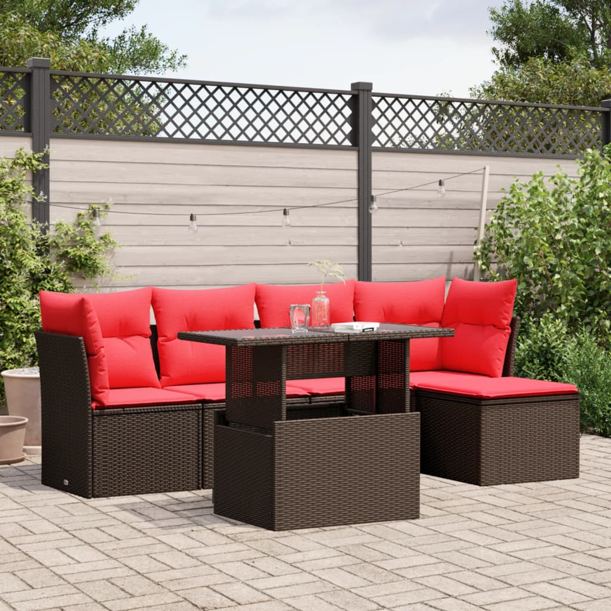 Terrassenmöbel-Set mit roten Kissen. Braune Korbmöbel, Glastisch und Hintergrund aus Holzzaun und Grün.