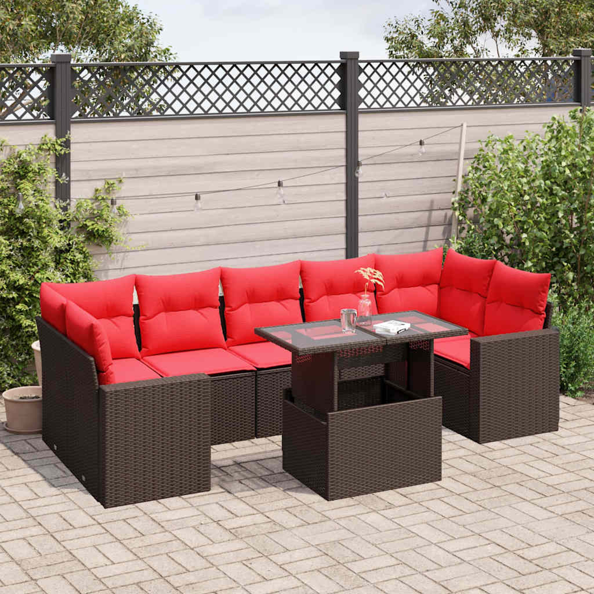 Outdoor-Wicker-Ecksofa mit roten Kissen und Glastisch auf einer Ziegelterrasse. Zaun und Grün im Hintergrund.